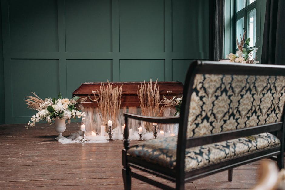 A close-up view of a floral arrangement placed on a wooden floor next to a vintage upholstered sofa with a floral pattern in a softly lit room. The arrangement features a large white ceramic vase fill