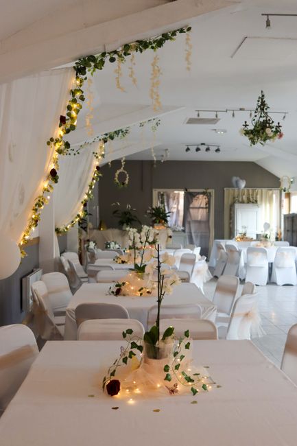 Interior view of a decorated event venue featuring a long, white-draped table with floral centerpieces composed of white roses, greenery, and small white flowers, surrounded by white chairs. The venue