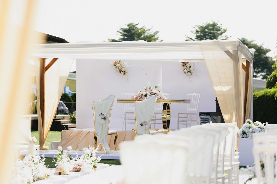 A floral arrangement scene at an outdoor wedding venue with a stage decorated with pastel pink and white flowers, including roses and peonies, arranged in a cascading style. The stage features a white
