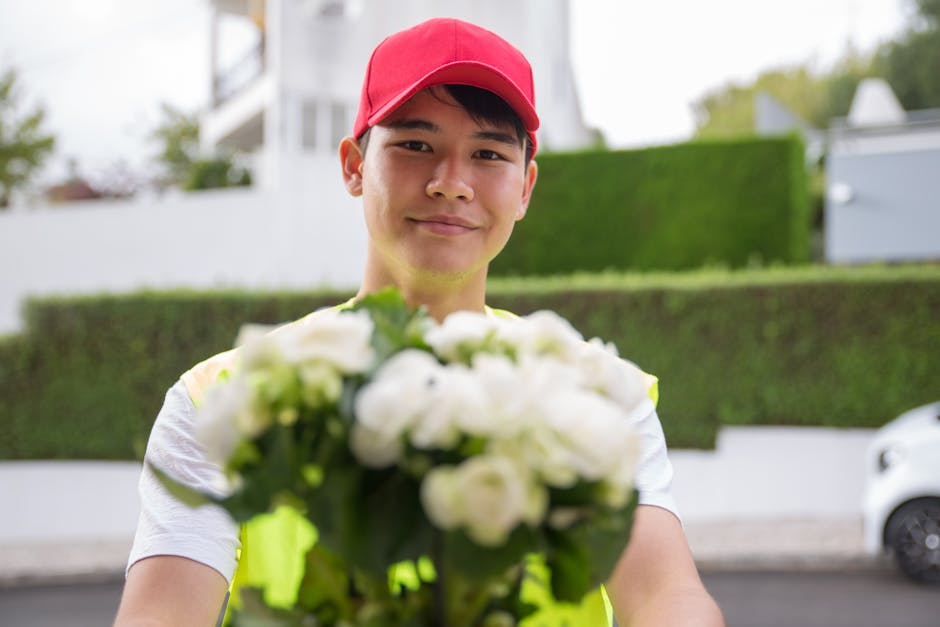 A delivery person wearing a red cap and light-colored T-shirt extending an arm forward, holding a bouquet of white flowers with green foliage, wrapped in clear cellophane. The background shows a resid