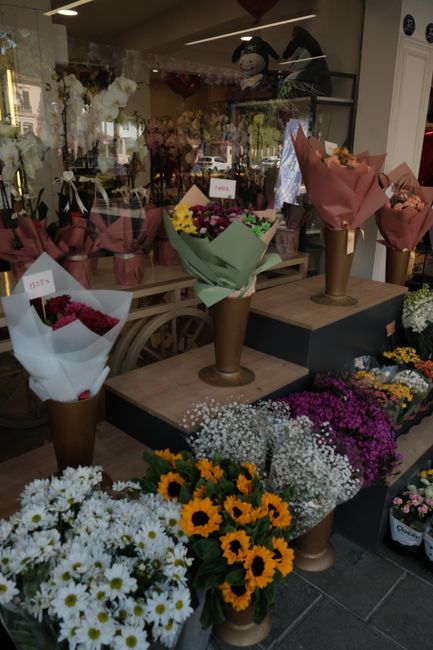 A display of fresh flower bouquets arranged on a wooden table inside a flower shop, featuring a variety of colorful floral gifts. The central bouquets are wrapped in pastel pink, green, and white pape