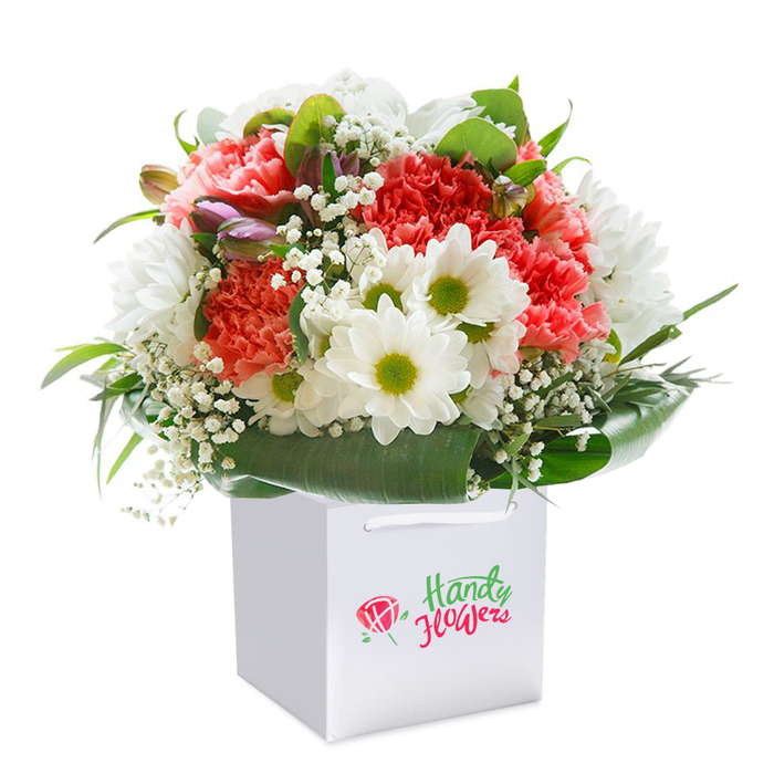 A radiant floral arrangement bursts to life within a clean, square white gift bag, set centre stage against a flawless white backdrop-highlighting every hue, texture, and detail. The bouquet's main feature is an abundance of pastel coral and lively red carnations, their layered, frilly petals creating a tapestry of softness and warmth, reminiscent of blooms found along the winding paths through Leyton Jubilee Park. Among the carnations, daisy-like white flowers catch the eye, their vibrant lime-green centres shining with a quiet cheer, much like the optimism radiating from the neighbourhood on a sunny afternoon. Wispy sprays of baby's breath thread through the denser flowers, bringing an element of grace and breathability, while subtle nods of purple-tinged buds-perhaps alstroemeria on the verge of opening-suggest the ever-changing nature of a local florist's daily selection, inspired by Leyton's cultural mosaic. Surrounding the entire display, lush, glossy green leaves form a natural base and elegant wrap, their sheen adding structure and a touch of garden-fresh luxury. The white gift bag, fitted with crisp ribbon handles, is designed for both charm and convenience, ideal for easy delivery to homes along Leyton's bustling avenues. Every floral element is thoughtfully placed, composed with the know-how only a neighbourhood expert can provide; the finished effect is one of generosity, freshness, and sincere celebration-an ideal gift for marking birthdays, anniversaries, or caring gestures in Leyton's E10 community.