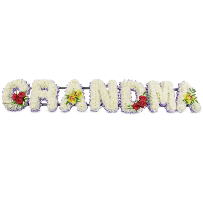 A detailed sympathy arrangement in the photograph spells out the word "GRANDMA" in large, ornate capitals, the letters arranged in a straight line as though resting on a stand in a chapel or crematorium. Each letter is intricately formed from tightly packed white chrysanthemums, giving a dense, softly rounded texture that looks both plush and refined. Underneath the white blooms, a slim band of pale lavender flowers peeks out at the base of every letter, creating a subtle outline that gently separates each shape from the bright white background. The 'G' is thoughtfully decorated with three deep red roses snuggled into the lower curve of the letter, their velvety petals and dark greenery adding a strong, emotional note. The first 'A' features two yellow-green orchids, their elongated petals and speckled centres lending a delicate, exotic touch that feels very personal. The letters 'R', 'N' and 'D' are kept purely floral in white, with only their lavender edging, which reinforces a sense of simplicity and peace. The 'M' repeats the motif of a small cluster of red roses tucked into the lower right, visually linking back to the 'G', while the final 'A' mirrors the first 'A' with another pair of fresh green orchids. A dark metal support frame runs along the back of the arrangement, holding the letters together as a single, stable piece that could be displayed at services in Leyton, nearby churches, or cemeteries along local routes. Overall, the tribute feels like a tender, hand-crafted message of gratitude, honouring a grandmother's loving role within the family and the community.